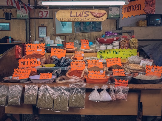 A market stall displaying a variety of spices and dried goods. Brightly colored signs with handwritten labels indicate different spices and mixtures, such as merk&eacute;n, cumin, and salt blends. The setup includes baskets, plastic containers, and plastic bags filled with goods, all organized neatly on a wooden counter. The signage also prominently features the name 'Luis, El casero del merken'.