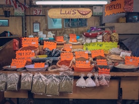 A market stall displaying a variety of spices and dried goods. Brightly colored signs with handwritten labels indicate different spices and mixtures, such as merkén, cumin, and salt blends. The setup includes baskets, plastic containers, and plastic bags filled with goods, all organized neatly on a wooden counter. The signage also prominently features the name 'Luis, El casero del merken'.