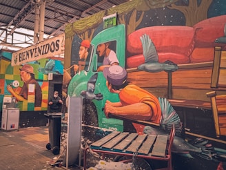 A mural on a wall depicting a vibrant scene with three people riding in a green truck, surrounded by birds. The mural shows a driver and two passengers in a dynamic pose. Below the mural, there is a person wearing a mask beside a garbage bin. The upper part of the mural has trees, and there are various objects like a bench, a fire extinguisher, and bins in the immediate surroundings. A sign reading 'BIENVENIDOS' (Welcome) is painted on a different wall in the background.