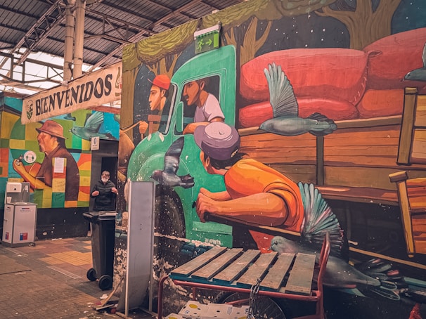 A mural on a wall depicting a vibrant scene with three people riding in a green truck, surrounded by birds. The mural shows a driver and two passengers in a dynamic pose. Below the mural, there is a person wearing a mask beside a garbage bin. The upper part of the mural has trees, and there are various objects like a bench, a fire extinguisher, and bins in the immediate surroundings. A sign reading 'BIENVENIDOS' (Welcome) is painted on a different wall in the background.