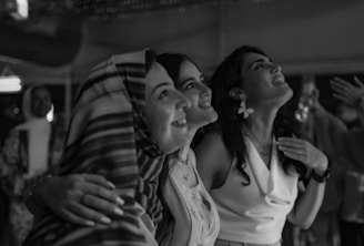 Three people are closely huddled together, smiling and looking upwards. The environment suggests a warm, indoor setting with soft lighting. They are capturing a joyful moment, possibly during a celebration or gathering.