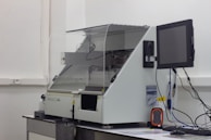 A laboratory machine with a protective transparent cover is positioned on a counter. Next to it, a monitor is attached, and various cables are connected. The setting appears to be sterile, with a focus on technology and instrumentation.