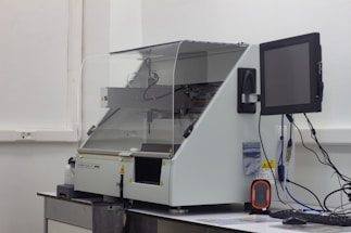 A laboratory machine with a protective transparent cover is positioned on a counter. Next to it, a monitor is attached, and various cables are connected. The setting appears to be sterile, with a focus on technology and instrumentation.