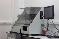 A laboratory machine with a protective transparent cover is positioned on a counter. Next to it, a monitor is attached, and various cables are connected. The setting appears to be sterile, with a focus on technology and instrumentation.