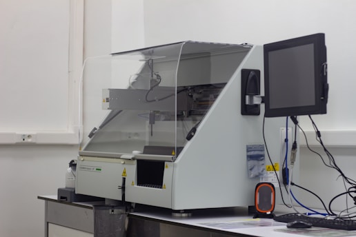 A laboratory machine with a protective transparent cover is positioned on a counter. Next to it, a monitor is attached, and various cables are connected. The setting appears to be sterile, with a focus on technology and instrumentation.