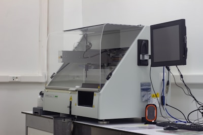 A laboratory machine with a protective transparent cover is positioned on a counter. Next to it, a monitor is attached, and various cables are connected. The setting appears to be sterile, with a focus on technology and instrumentation.