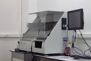 A laboratory machine with a protective transparent cover is positioned on a counter. Next to it, a monitor is attached, and various cables are connected. The setting appears to be sterile, with a focus on technology and instrumentation.