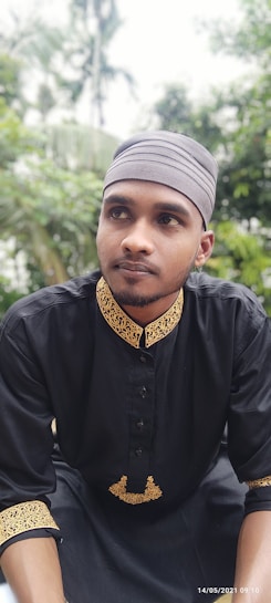 A person is wearing a black traditional outfit with intricate gold embroidery. The attire includes a matching head covering in a gray shade. The background features lush greenery, suggesting an outdoor setting with trees and blurred foliage.