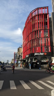 A street view featuring a modern multi-story building with a striking red facade. The building is a Gloria Jean's Coffees shop, identifiable by the signage. Several motorcycles are on the road, with a cyclist in the foreground. The sky is partly cloudy, and the overall scene captures an urban environment.