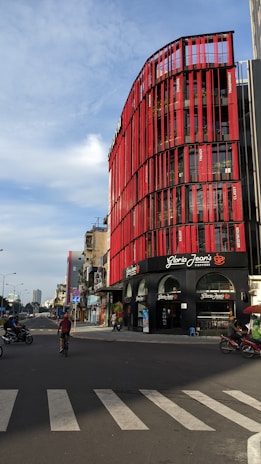 A street view featuring a modern multi-story building with a striking red facade. The building is a Gloria Jean's Coffees shop, identifiable by the signage. Several motorcycles are on the road, with a cyclist in the foreground. The sky is partly cloudy, and the overall scene captures an urban environment.