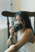 A woman with long dark hair is sitting in a chair by a window, holding a vintage-style camera close to her face. The soft lighting and pastel butterfly decorations on the window create a serene atmosphere.