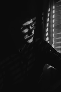 A person stands in a dark space with slits of light from window blinds casting dramatic shadows across their face and body. The angle of the light creates a pattern of alternating light and shadow, highlighting the contemplative expression on their face.