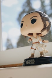 A Star Wars-themed vinyl figurine is displayed on a windowsill. The character is dressed in white attire with brown boots and a black belt. The figure stands on a black base that has 'Star Wars' written on the front. Sunlight filters through a netted window in the background, creating a bright and airy setting.