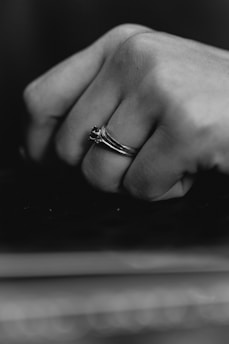 A close-up black and white photograph of a hand with an elegant ring on the ring finger, showing a minimalist design with a dark stone.