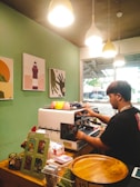 A person operates a coffee machine in a cozy café corner with green walls adorned with artwork. Pendant lights hang from the ceiling, illuminating the workspace. Various items like jars, a wooden tray, and printed materials are neatly arranged on the counter. Natural light streams in through a window, providing a glimpse of the outdoors.