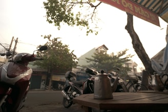 An outdoor scene featuring several parked motorcycles in front of a tree-lined street. A metal kettle is placed on a wooden table in the foreground, with residential buildings visible in the background. Overhead, a partially visible sign and a tree with sparse leaves can be seen against a bright sky.