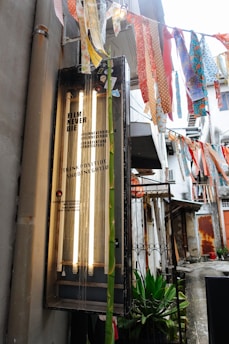 An outdoor alleyway features a retro-styled lightbox with the text 'Film Never Die' and several colorful fabric strips hanging above. The scene includes a hint of greenery and aged urban architecture in the background.