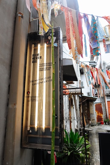An outdoor alleyway features a retro-styled lightbox with the text 'Film Never Die' and several colorful fabric strips hanging above. The scene includes a hint of greenery and aged urban architecture in the background.