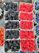 A variety of fresh berries arranged in cartons, including blackberries, raspberries, strawberries, and blueberries. Each type is neatly organized in individual compartments on a table. The vibrant colors of the berries create an appealing visual display.