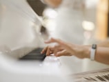 Hands are playing a white piano, with the focus on the fingers pressing the keys. The background is softly blurred, suggesting an elegant and serene setting.
