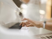 Hands are playing a white piano, with the focus on the fingers pressing the keys. The background is softly blurred, suggesting an elegant and serene setting.