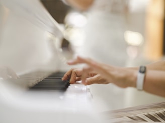 Hands are playing a white piano, with the focus on the fingers pressing the keys. The background is softly blurred, suggesting an elegant and serene setting.