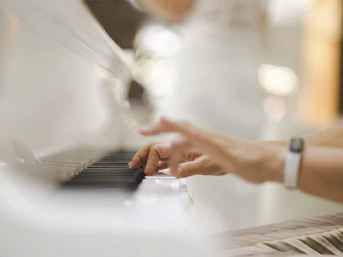 Hands are playing a white piano, with the focus on the fingers pressing the keys. The background is softly blurred, suggesting an elegant and serene setting.