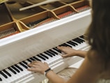 Hands are playing on a white wooden piano, with visible strings and hammers inside the instrument.