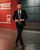 A man in a tailored black suit walks confidently in an urban setting. He is wearing a gray waistcoat, a black tie, and black polished shoes. His hair is slicked back, and he has a well-groomed beard. Behind him is a red wall with geometric designs and an arrow symbol.