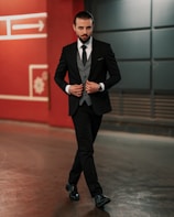 A man in a tailored black suit walks confidently in an urban setting. He is wearing a gray waistcoat, a black tie, and black polished shoes. His hair is slicked back, and he has a well-groomed beard. Behind him is a red wall with geometric designs and an arrow symbol.