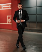A man in a tailored black suit walks confidently in an urban setting. He is wearing a gray waistcoat, a black tie, and black polished shoes. His hair is slicked back, and he has a well-groomed beard. Behind him is a red wall with geometric designs and an arrow symbol.