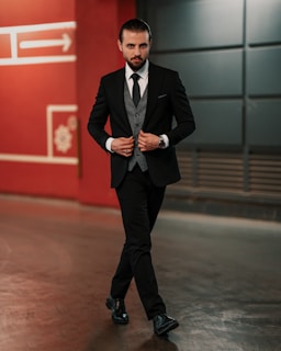 A man in a tailored black suit walks confidently in an urban setting. He is wearing a gray waistcoat, a black tie, and black polished shoes. His hair is slicked back, and he has a well-groomed beard. Behind him is a red wall with geometric designs and an arrow symbol.
