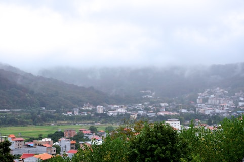 A small town nestled at the foot of lush, green hills covered in mist. The town features a mix of residential buildings with colorful roofs, surrounded by dense tree foliage. The sky is overcast, giving the scene a serene and tranquil vibe.