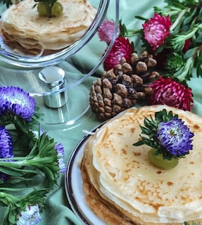 A stack of crepes garnished with a green grape and a purple flower sits on a plate. Surrounding the plate are vibrant flowers and pine cones placed on a green fabric background, creating a visually appealing and rustic presentation.