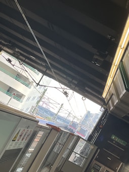 A train station platform featuring overhead cables and infrastructure is visible. There are trains in the distance, with some visible signage on the platform. The scene is framed by the structural elements of the station, including wires, poles, and a partial view of a roof.