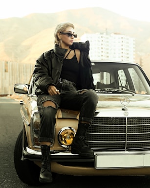A person with short hair, wearing sunglasses, a black top, and a black puffy jacket sits on the hood of a vintage beige Mercedes in front of a mountainous landscape and urban buildings. They wear ripped jeans and black boots, exuding a stylish, rebellious look.