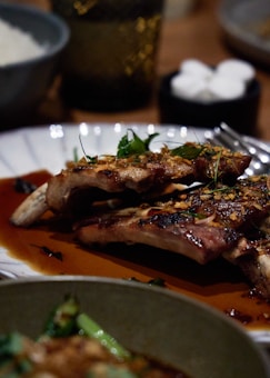 A close-up view of a plate with grilled ribs garnished with herbs, sitting in a rich brown sauce. The background includes a blurred dish of white rice and some out-of-focus drinking glasses.