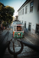 A colorful tram covered with graffiti ascends a steep cobblestone street between historic buildings. The tram is approaching from the distance, framed by large trees on one side and a wall with urban graffiti on the other. There are stairs in the foreground with iron railings.