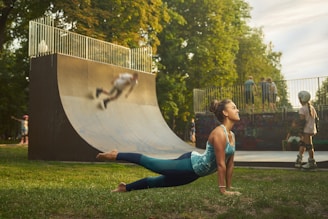 A woman is practicing yoga on grass in a park, performing a cobra pose. Nearby, children are playing on skateboards and rollerblades around a half-pipe or skate ramp, with one skater blurred in motion. Trees with lush green foliage surround the area, giving a peaceful outdoor atmosphere.