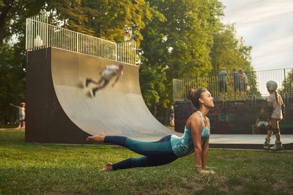 A woman is practicing yoga on grass in a park, performing a cobra pose. Nearby, children are playing on skateboards and rollerblades around a half-pipe or skate ramp, with one skater blurred in motion. Trees with lush green foliage surround the area, giving a peaceful outdoor atmosphere.