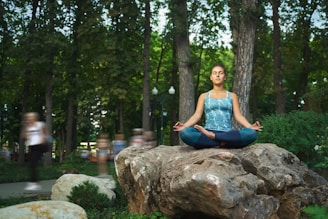 A person sits cross-legged in a meditative pose on a large rock, surrounded by lush trees in a park. The individual wears a blue patterned tank top and leggings, with eyes closed, exuding a sense of calm amidst the blurred motion of passersby in the background.