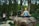 A person sits cross-legged in a meditative pose on a large rock, surrounded by lush trees in a park. The individual wears a blue patterned tank top and leggings, with eyes closed, exuding a sense of calm amidst the blurred motion of passersby in the background.