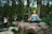 A person sits cross-legged in a meditative pose on a large rock, surrounded by lush trees in a park. The individual wears a blue patterned tank top and leggings, with eyes closed, exuding a sense of calm amidst the blurred motion of passersby in the background.