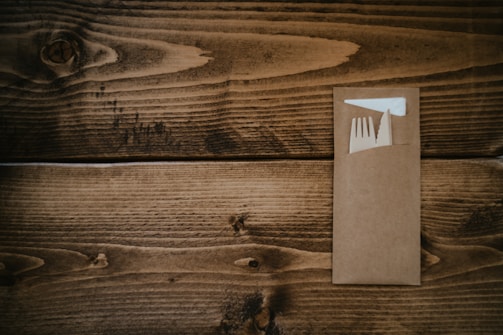 A wooden surface with a brown envelope containing a plastic fork and a white napkin.