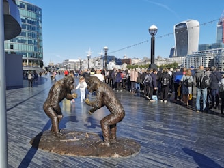 A pair of gorilla statues made of bronze stand face-to-face on a large urban promenade. Numerous pedestrians, many with backpacks and casual attire, walk and gather along the promenade. Modern high-rise buildings, including distinctive glass and curved architectural designs, form the background. The bright blue sky indicates a clear and sunny day.