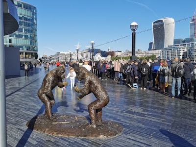 A pair of gorilla statues made of bronze stand face-to-face on a large urban promenade. Numerous pedestrians, many with backpacks and casual attire, walk and gather along the promenade. Modern high-rise buildings, including distinctive glass and curved architectural designs, form the background. The bright blue sky indicates a clear and sunny day.
