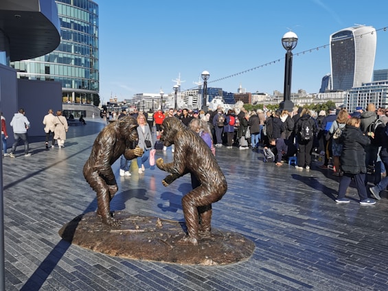Two bronze statues of gorillas engaging in a playful interaction are prominently displayed on a modern city promenade. The scene is bustling with people walking and gathering in the background. Buildings and skyscrapers with unique architectural designs are visible under a clear blue sky.