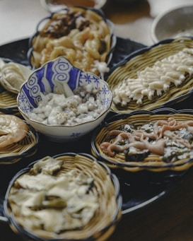 A selection of dishes is presented in small woven baskets on a dark platter. The central dish is in a blue and white patterned bowl filled with a creamy white substance, possibly a salad or dessert. Surrounding the bowl are various traditional dishes with different textures and colors, including noodles, greens, and seafood.