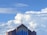 A church building with a large cross on top, set against a backdrop of dramatic, fluffy clouds and a bright blue sky. The structure features distinctive blue stained-glass windows and a brown brick facade, contributing to its ornate architectural style.