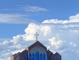 A church building with a large cross on top, set against a backdrop of dramatic, fluffy clouds and a bright blue sky. The structure features distinctive blue stained-glass windows and a brown brick facade, contributing to its ornate architectural style.